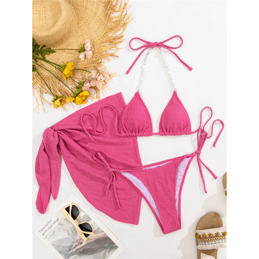 Alluring three-piece bikini set for women featuring delicate pearl embellishments and a textured, wrinkled design. Includes a sarong and halter top made of Nylon and Spandex. Comes with a pad and offers a low waist, wire free support. The bikini set fits true to size and is available in sizes S, M, and L. Colors available are hot pink, blue, purple, and white. Free shipping offered on this versatile beach-to-bistro style swimwear. Ideal for women aged 18 to 35.